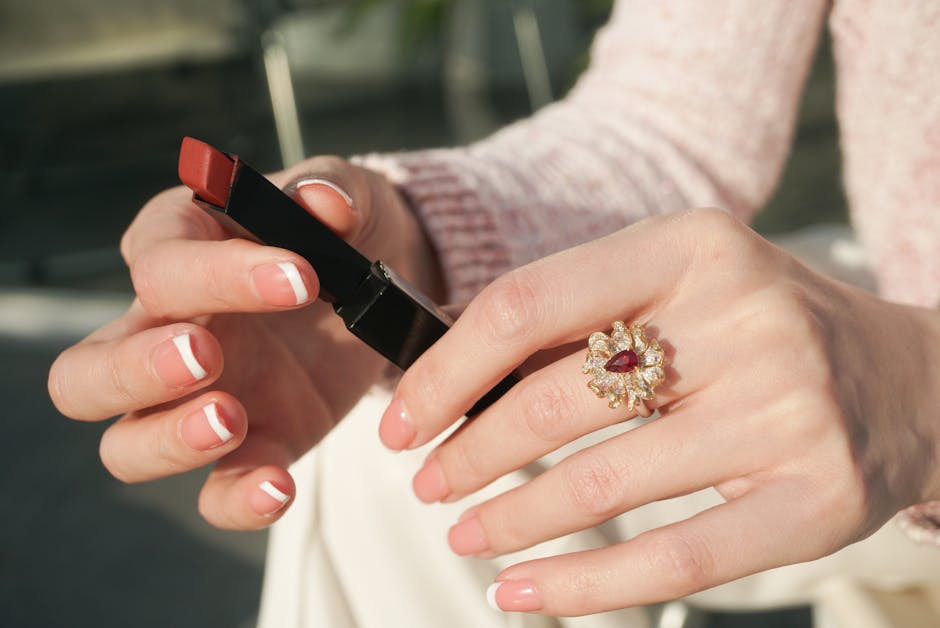 Elegant hands with French manicure holding lipstick, featuring a ruby ring in a sunny setting.