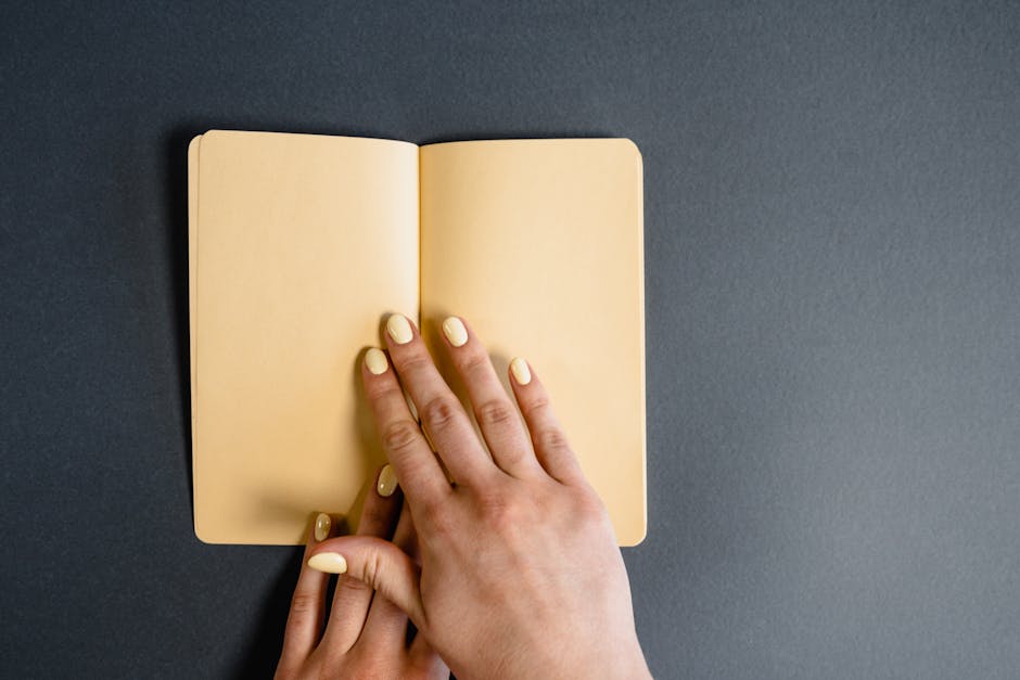 Close-up of hands with yellow nails holding an open yellow notebook on a grey background.
