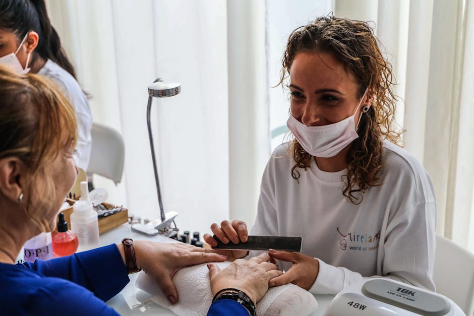 A beautician in a mask manicures a client's nails in a bright salon.